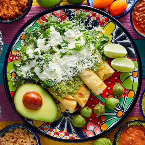 A colorful plate featuring enchiladas topped with green sauce, cheese, lettuce, avocado, and lime, surrounded by various sides.