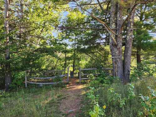 A wooded path leads through trees to a clearing, with a wooden fence and a sign visible in the background.