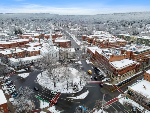 Aerial view of a snowy town square with buildings, trees, and a circular road surrounded by snow-covered hills.