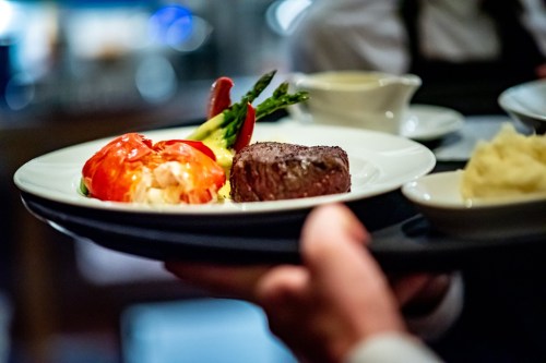 A waiter serves a plate with lobster, steak, asparagus, and mashed potatoes in a fine dining setting.