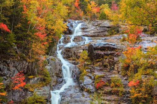 A cascading waterfall flows down rocky cliffs, surrounded by vibrant autumn foliage in shades of orange and yellow.