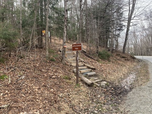 A wooden sign marks the Appalachian Trail, with steps leading into a wooded area beside a gravel road.
