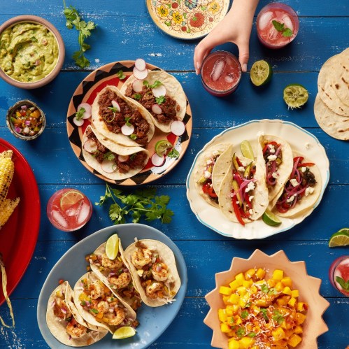 A vibrant spread of tacos, guacamole, and drinks on colorful plates, garnished with fresh herbs and lime.