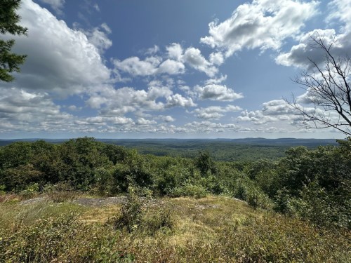 A panoramic view of lush green hills under a partly cloudy sky, with distant mountains visible on the horizon.