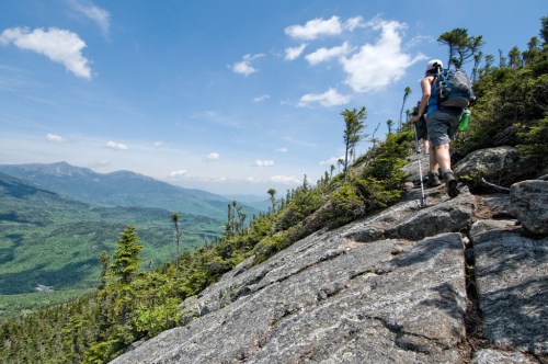 A hiker ascends a rocky trail with a scenic view of green mountains and blue skies in the background.