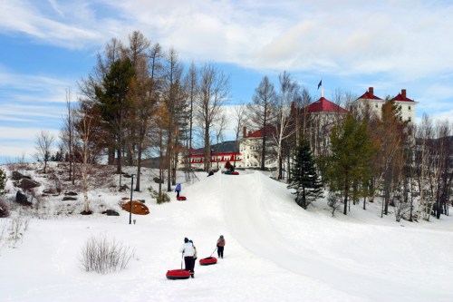 Two people pull sleds up a snowy hill, with a historic building and trees in the background under a blue sky.