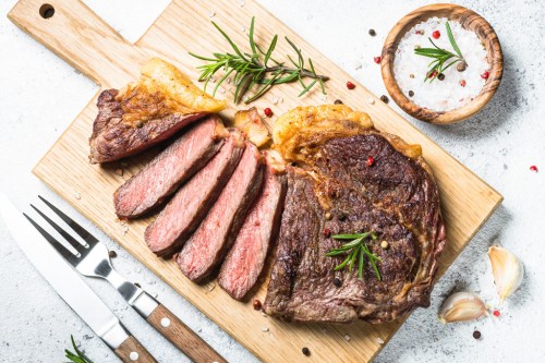 A wooden cutting board with a sliced steak, garnished with rosemary and pepper, alongside a bowl of salt and garlic.