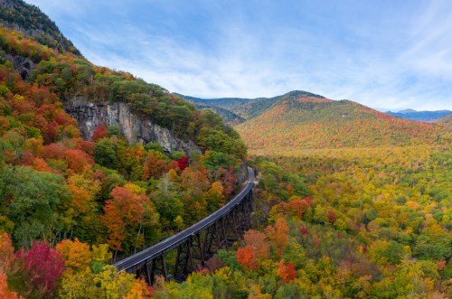 A scenic view of a winding railway through vibrant autumn foliage and mountains under a blue sky.