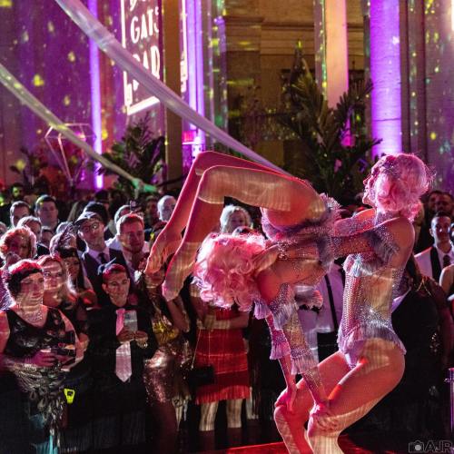 A lively performance with two dancers in sparkly outfits, surrounded by an audience in a colorful, festive setting.