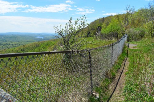 A chain-link fence along a path overlooking a lush green valley and distant hills under a blue sky.