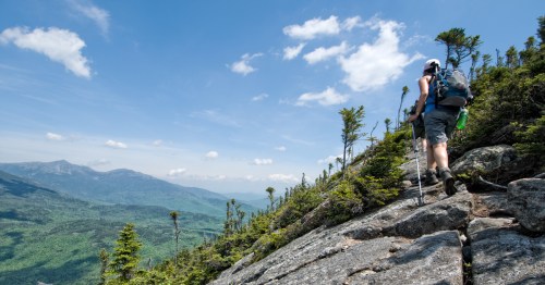 A hiker ascends a rocky trail with lush green mountains and a blue sky in the background.
