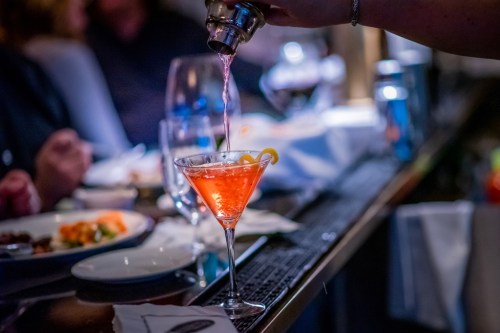 A cocktail being poured into a glass, with a lemon twist, set against a blurred dining scene.