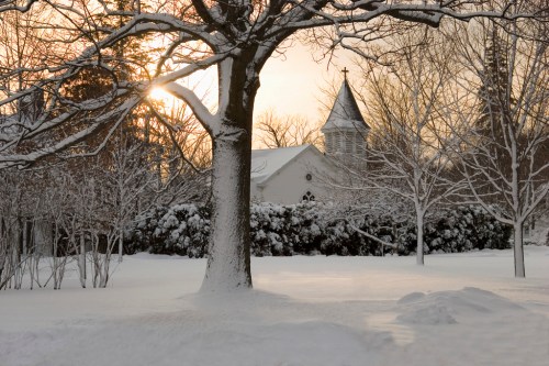 A snowy landscape with a church in the background, framed by bare trees and a soft sunset glow.