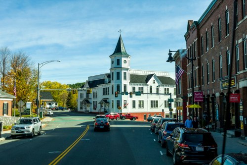 A quaint town square featuring a historic white building with a tower, surrounded by shops and parked cars.