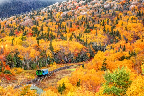 A vintage train travels along a winding track through vibrant autumn foliage in a mountainous landscape.