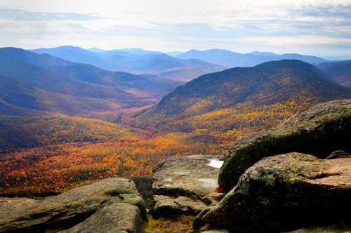A panoramic view of colorful autumn foliage in a mountainous landscape under a cloudy sky.