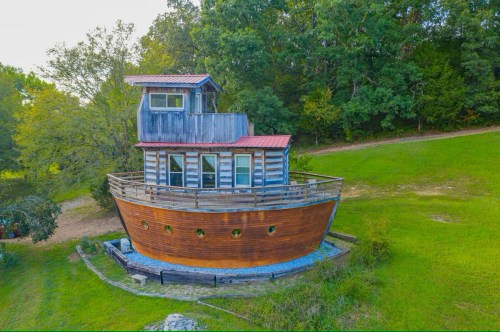 A unique wooden house designed to resemble a boat, set on a grassy hillside surrounded by trees.
