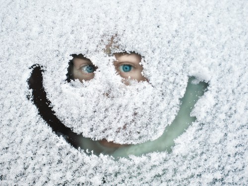 A child's face peeks through a snow-covered window, with a smiley face shape cleared in the snow.