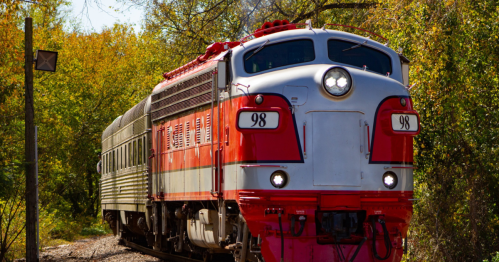 A vintage red and silver train travels along a track surrounded by lush green trees.