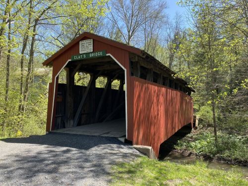 A red covered bridge named Clay's Bridge surrounded by lush greenery and trees on a sunny day.