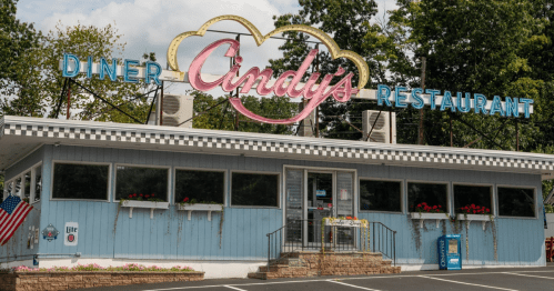 A blue diner with a pink sign reading "Cindy's Restaurant," featuring outdoor seating and American flags.