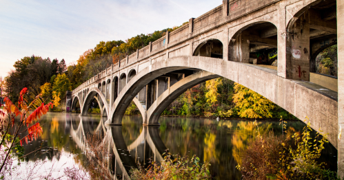 A concrete arch bridge spans a calm river, surrounded by colorful autumn foliage reflecting in the water.