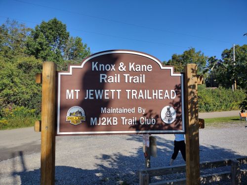 Sign for Mt. Jewett Trailhead on Knox & Kane Rail Trail, maintained by MJ2KB Trail Club, with a clear blue sky.