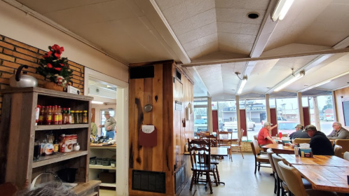 Cozy diner interior with wooden decor, shelves of condiments, and patrons enjoying meals at tables.