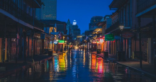 A quiet street at night, illuminated by colorful neon lights reflecting on wet pavement in a city.