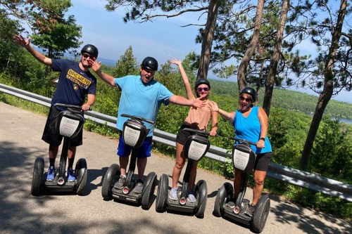 Four people on segways, smiling and posing with arms raised, surrounded by trees and a scenic landscape.