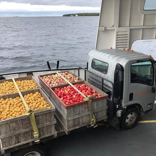 A truck on a ferry transporting wooden crates filled with various fruits, with a scenic water view in the background.