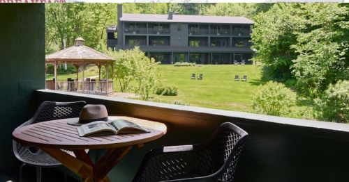 A balcony view featuring a wooden table with a hat and magazine, overlooking a green lawn and a gazebo.