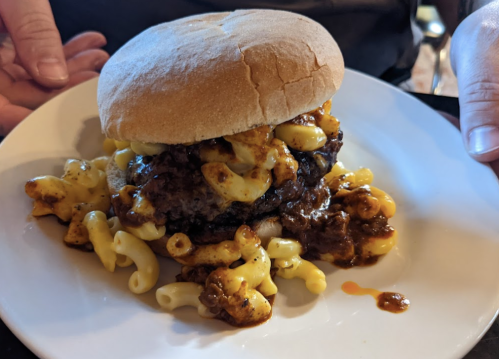 A hamburger topped with macaroni and cheese, served on a plate with a soft bun.