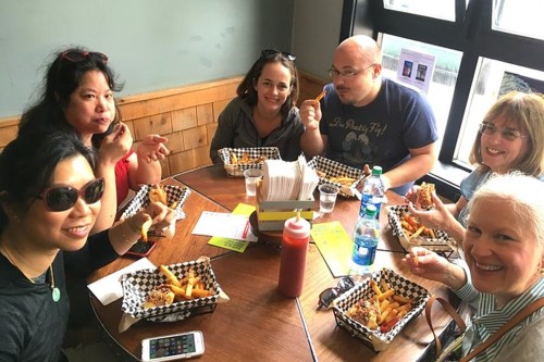 A group of six people enjoying food at a table, with fries and drinks, smiling and holding their meals.