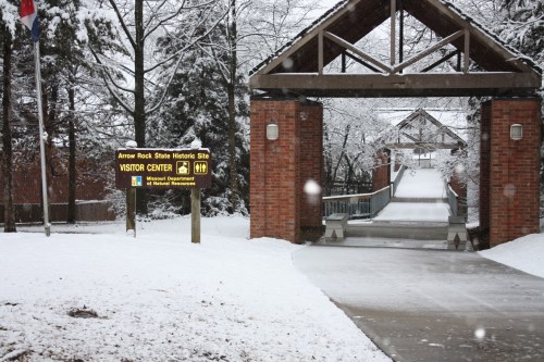Snow-covered entrance to the Visitor Center at Arrow Rock State Historic Site, with a sign and bridge in view.