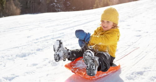 A child in a yellow jacket and hat joyfully sleds down a snowy hill, smiling with excitement.