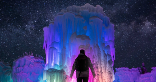 A person stands before colorful ice formations under a starry night sky.