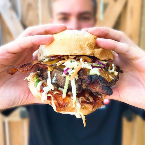 A person holds a loaded burger with bacon, coleslaw, and barbecue sauce, against a wooden background.