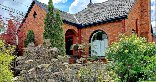 A charming brick house with a landscaped garden featuring green shrubs and blooming flowers under a blue sky.