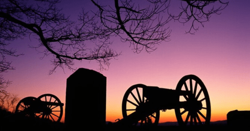 Silhouetted cannons against a vibrant sunset, with bare branches in the foreground.