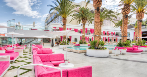 A vibrant pool area with pink lounge chairs, palm trees, and a modern building in the background under a blue sky.