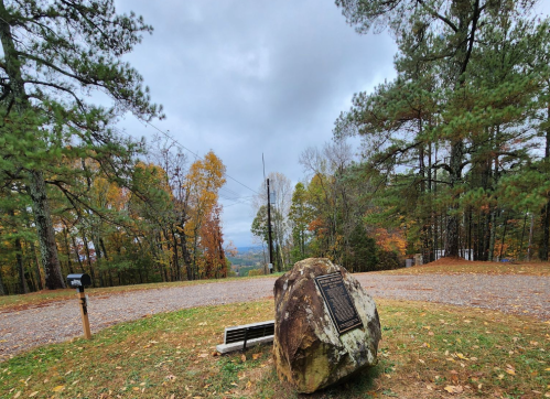 A scenic view from a hillside with a large rock and plaque, surrounded by trees and autumn foliage under a cloudy sky.