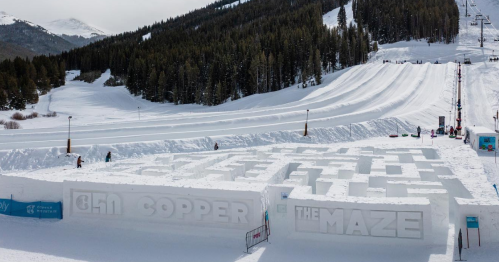 A snowy maze structure in a winter landscape, surrounded by trees and ski slopes.