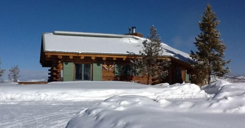 A log cabin covered in snow, surrounded by trees under a clear blue sky.