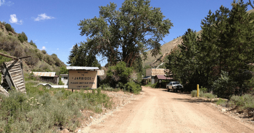 Dirt road leading to a sign for "J-Bridge," surrounded by trees and abandoned buildings under a blue sky.