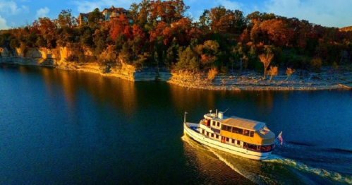 A boat glides across a calm lake, surrounded by autumn-colored trees and rocky cliffs under a clear blue sky.