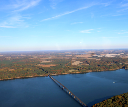 Aerial view of a river with a bridge, surrounded by lush green fields and a clear blue sky.