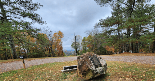 A scenic view from a hilltop with a plaque on a rock, surrounded by autumn trees and a cloudy sky.