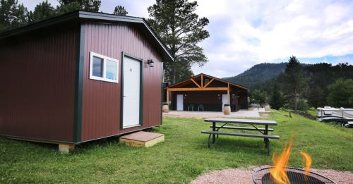 A brown cabin and a larger building in a scenic outdoor setting with a picnic table and a fire pit.