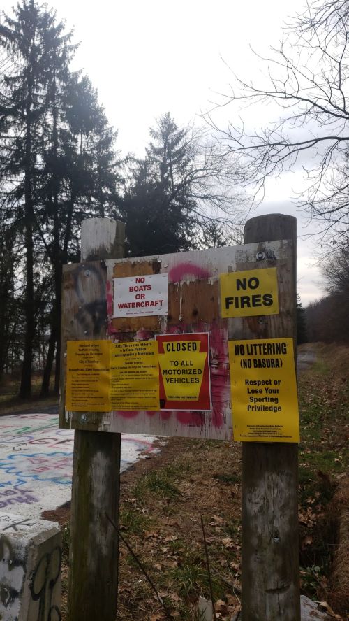 A weathered sign with multiple warnings: no boats, no fires, closed to motorized vehicles, and no littering. Trees in background.